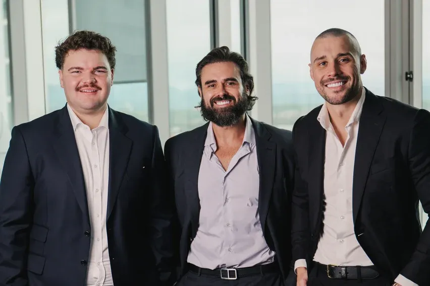 The Amassed team, from left: Lachlan Hodgson, Scott Richards and Elijah Turkovic, standing together in dark suits beside floor-to-ceiling windows.