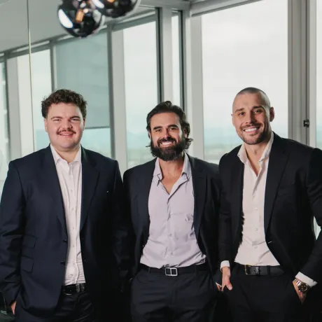 The Amassed team, Lachlan Hodgson, Scott Richards and Elijah Turkovic, standing together in dark suits beside floor-to-ceiling windows