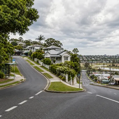 A forked road dividing between two contrasting Brisbane neighbourhoods under cloudy skies, representing the risks of buying in the wrong suburb and the importance of careful suburb selection.